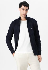Marine blå strikket cardigan med kabelmønster, ribbede mansjetter og krage; har glidelåsåpning og liten logo på brystet. Kombinert med en hvit skjorte.