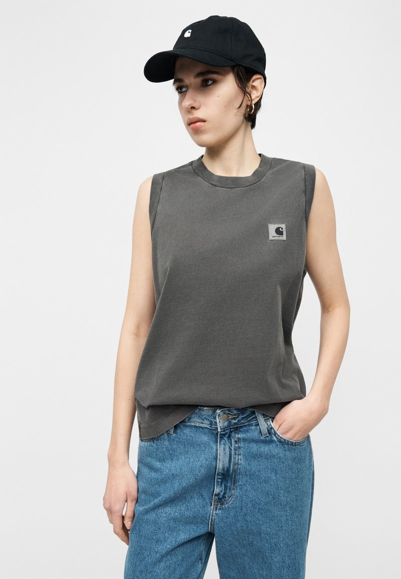 Person wearing a black cap, sleeveless dark gray shirt with a logo patch, and blue jeans, standing with one hand in pocket against a plain background.