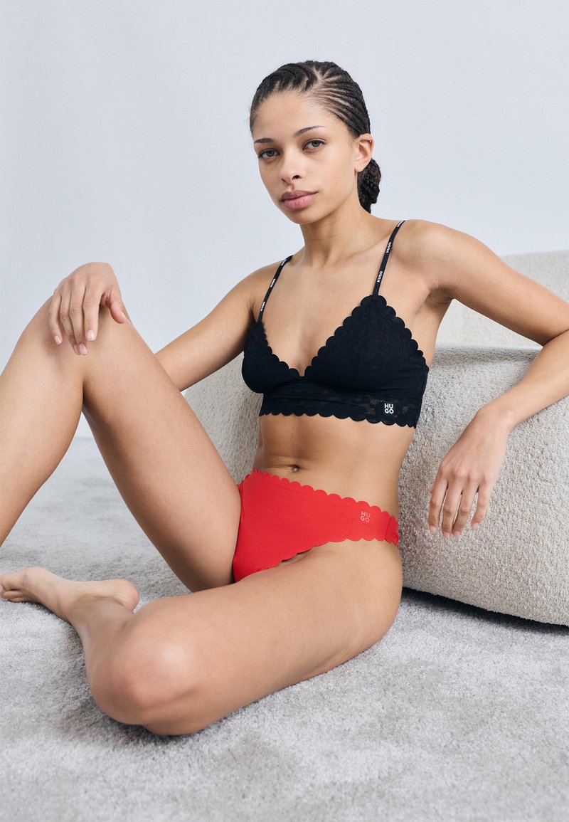 Bralette de encaje negro con bordes festoneados combinado con bragas rojas de talle alto con bordes festoneados. Modelo sentada sobre una superficie clara con textura.