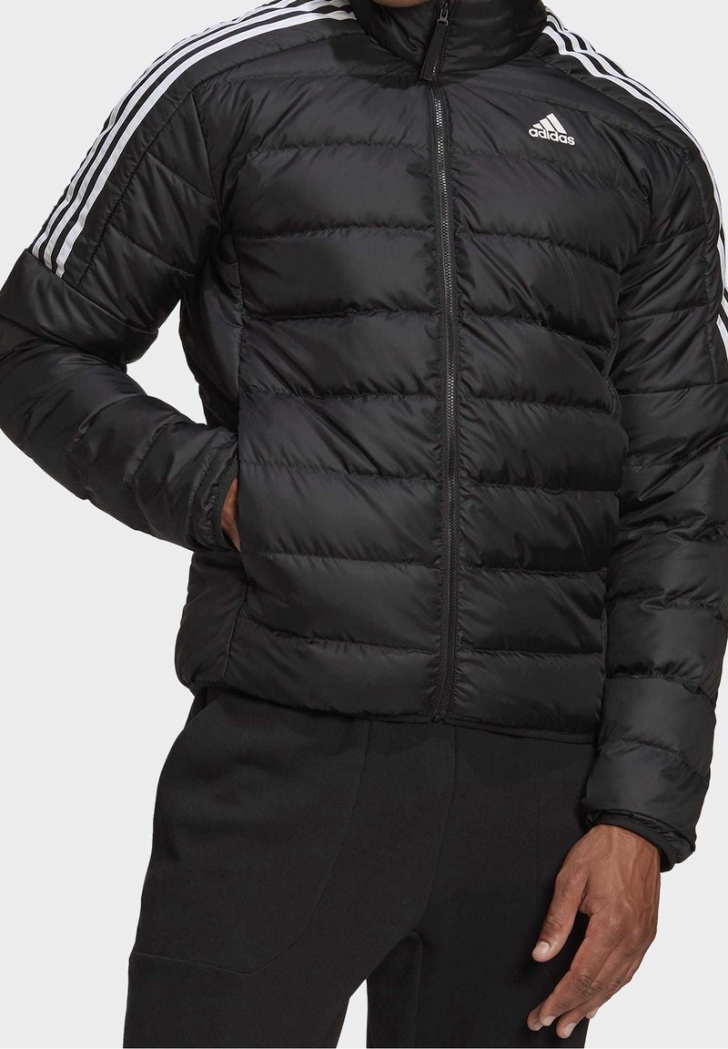 Doudoune noire Adidas avec des rayures blanches sur les manches et le logo sur la poitrine, portée avec le zip fermé, la main dans la poche gauche.