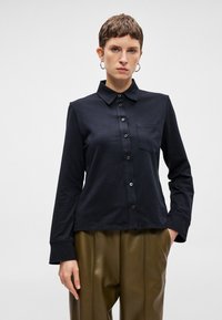 Črna srajca z dolgimi rokavi in ovratnikom, ki ima sprednje zapiranje na gumb in en sam žep na prsih. Gladek, mehak material z oblikovanim prilegom.