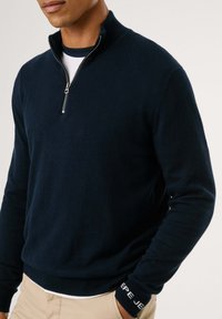 Maglione blu navy realizzato in materiale morbido, dotato di un collo a zip quarti e una vita a coste con dettaglio logo sulla manica.