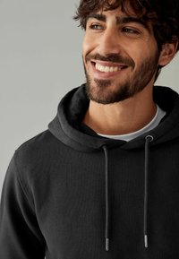 Black hoodie with a drawstring and silver tips, featuring a soft texture and kangaroo pocket, worn over a white shirt. Neutral background.