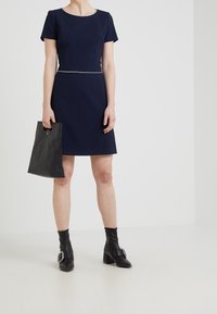 Robe trapèze bleu marine avec manches courtes, tissu texturé, bordure claire contrastante au niveau de l'encolure, associée à une pochette noire et des bottines.