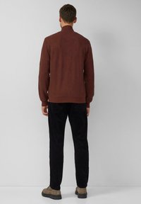 Brauner, strukturierter Pullover mit hohem Kragen, kombiniert mit schwarzen Cordhosen. Die Schuhe sind beige mit einer dicken schwarzen Sohle. Rückenansicht gezeigt.