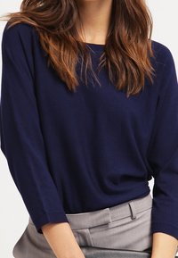 Námořnicky modrý lehký pletený svetr s třičtvrtinovými rukávy, lodičkovým výstřihem a uvolněným střihem, kombinovaný se šedými elegantními kalhotami.