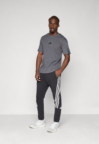 T-shirt de sport gris à manches courtes avec des accents noirs, associé à un jogging noir portant des rayures blanches sur les côtés et des baskets blanches.