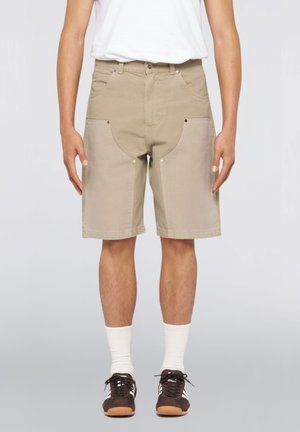 Persona che indossa pantaloncini beige fino al ginocchio con grandi tasche frontali, calzini bianchi, scarpe da ginnastica marrone scuro e una maglietta bianca, in piedi contro uno sfondo neutro.