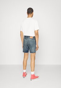 Hombre de espaldas con una camiseta blanca, pantalones cortos de mezclilla azules, calcetines blancos y zapatillas altas rosas contra un fondo blanco sencillo.