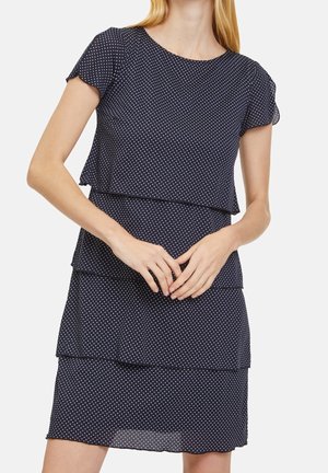 Mujer con vestido azul marino de manga corta con lunares blancos y detalles de volantes en capas, manos ligeramente entrelazadas al frente.