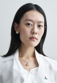 Goldene Ohrringe und eine passende Halskette mit einem kleinen Planetendesign, getragen mit einem weißen Shirt, das ein minimalistisch gesticktes Detail aufweist.