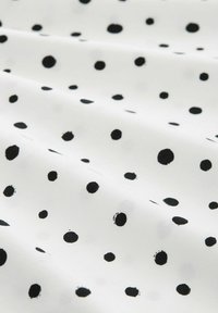 Weißer Stoff mit einem wiederholten Muster aus schwarzen Punkten in verschiedenen Größen. Die Textur wirkt glatt und weich, mit einem leichten Glanz.