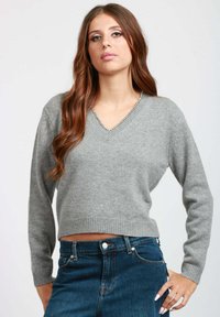 Giovane donna con lunghi capelli castani che indossa un maglione grigio a V e jeans blu a vita alta in posa con una mano sulla vita.