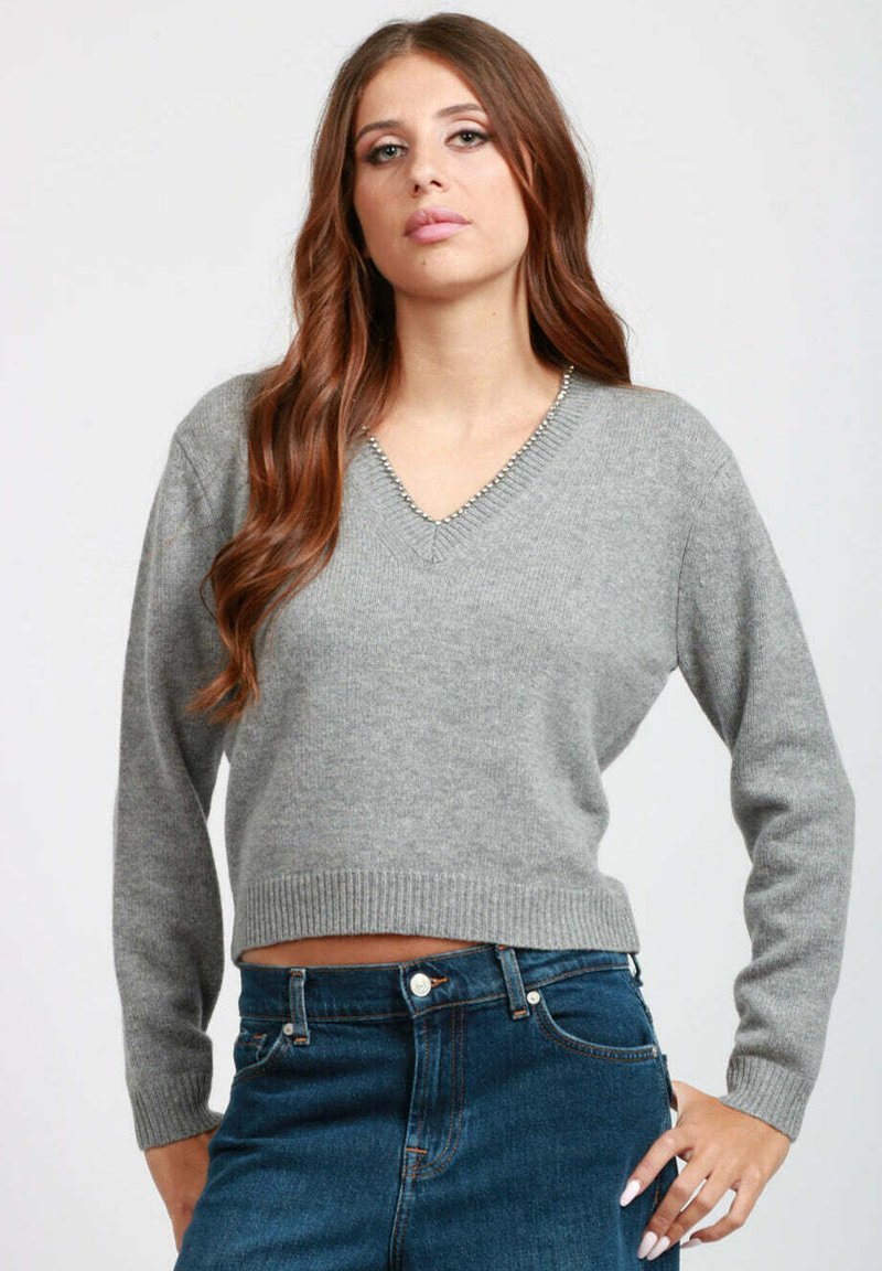 Giovane donna con lunghi capelli castani che indossa un maglione grigio a V e jeans blu a vita alta in posa con una mano sulla vita.