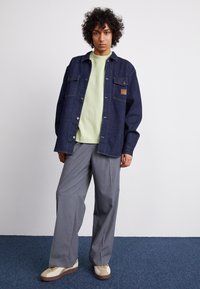 Denim jacket in dark blue with a chest pocket and branded patch, worn over a light green crewneck top. Paired with light grey wide-leg trousers and sneakers.