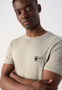 Gaiši pelēks T-krekls ar apaļu kakla izgriezumu; kreisajā krūtīs ir izšūti kafijas krūzes un vīna glāzes dizaini. Mīksta auduma tekstūra.