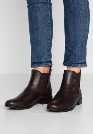 Femme portant des bottines Chelsea en cuir marron foncé avec des panneaux latéraux élastiques et un jean slim bleu.