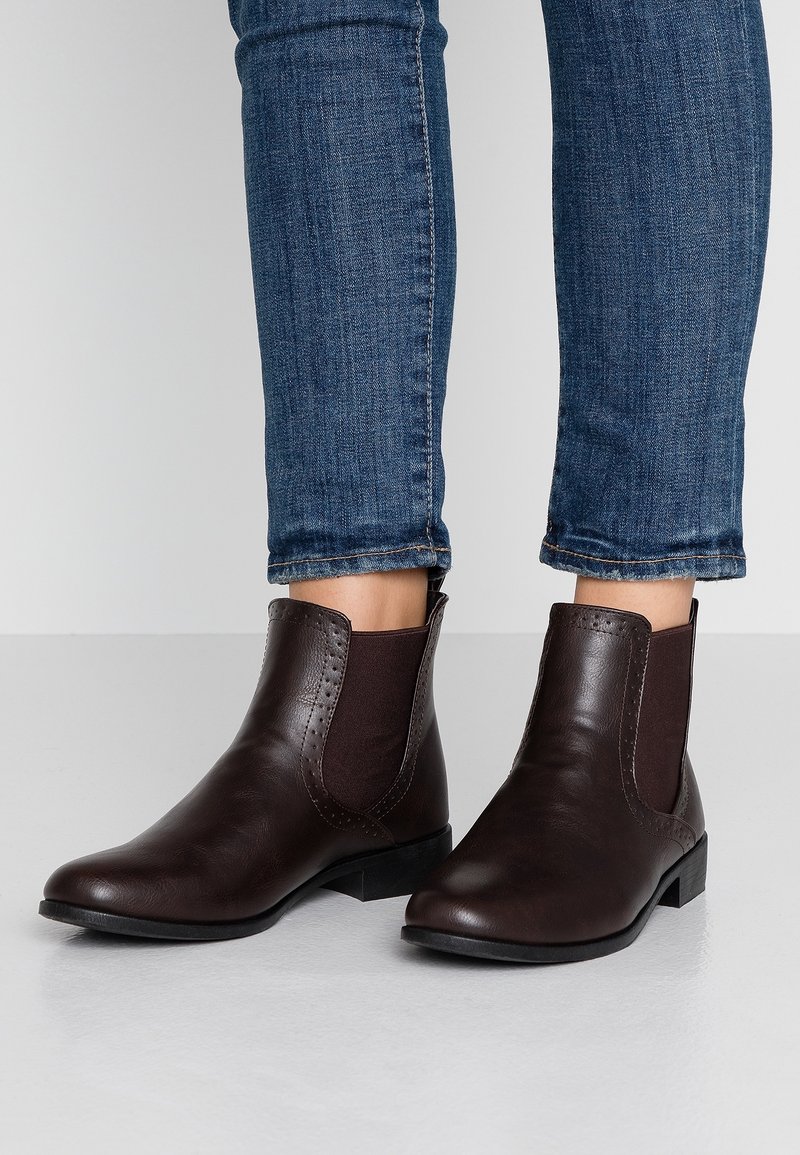Femme portant des bottines Chelsea en cuir marron foncé avec des panneaux latéraux élastiques et un jean slim bleu.