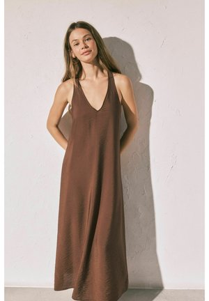 Jeune femme aux longs cheveux portant une robe longue marron sans manches, debout devant un mur clair texturé, les mains derrière le dos.