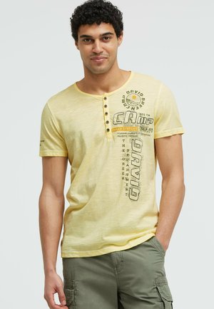Mann mit lockigem Haar trägt ein hellgelbes Henley-Shirt mit kurzen Ärmeln und grüne Cargo-Shorts, steht mit einer Hand in der Tasche.