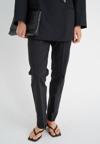 Mustahuolinen bleiseri ylle puettuna, sovitettujen mustien housujen kanssa, täydentäen ilmettä teksturoitulla mustalla laukun ja minimalistisilla hihnoilla varustetuilla mustilla flip-floppailla.