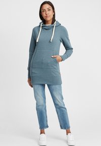 Vestido sudadera azul con cuello drapeado, cordones beige, bolsillo frontal y dobladillo acanalado. Combinado con jeans rectos de lavado claro y zapatos blancos.