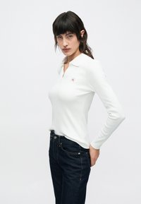 Biele polo tričko s dlhým rukávom, rebrovanou textúrou a malým červeným logom na hrudi, kombinované s tmavomodrými priliehavými džínsami.