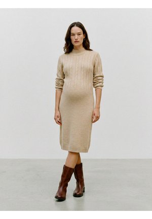 Donna che indossa un vestito beige a trecce lungo fino al ginocchio con maniche lunghe e stivali di pelle marroni a metà polpaccio, in piedi su un pavimento semplice.