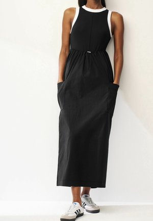Woman wearing a sleeveless black midi dress with white trim and pockets, paired with white and black Adidas sneakers, standing against a white wall.