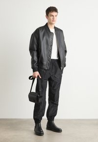 Schwarze Bomberjacke mit steppenden Details, weißes T-Shirt, schwarze strukturierte Jogginghosen und Chelsea-Stiefel, während er eine schwarze Umhängetasche hält.