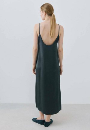 Donna con capelli biondi raccolti in una coda bassa, che indossa un vestito smanicato di raso nero con schiena scoperta e pantofole nere, in piedi su un pavimento grigio.