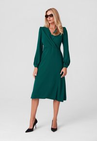 Zielona sukienka z długim rękawem, dekoltem w serek i marszczonymi ramionami; w kształcie litery A, gładka w dotyku, do kolan, noszona z czarnymi szpilkami.