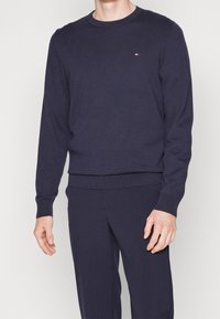 Granatowy sweter dzianinowy z długimi rękawami, okrągłym dekoltem i prążkowanym brzegiem. Posiada małe logo na lewej piersi. Miękka tekstura.