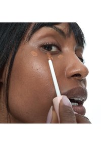 Femme appliquant un correcteur beige sous son œil droit avec un petit bâtonnet, se concentrant sur la peau près de l'os de la joue et la zone des yeux.