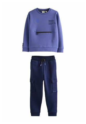 Lilla sweatshirt med en frontlomme med lynlås og sort lettering, parret med marineblå sweatpants med sidelommer og snor i taljen.