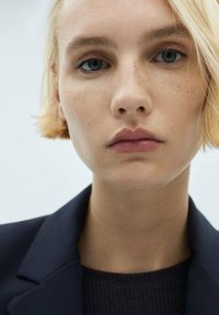 Primer plano de una persona con piel clara, cabello corto rubio y expresión neutra, vistiendo un blazer oscuro sobre una blusa texturizada.