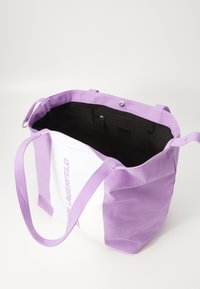 Lila och vit kanvas toteväska med långa handtag, öppen topp och svart inredning; har ett litet ficka med dragkedja och logotypdetalj på sidan.