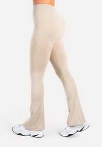 Beige uitlopende legging gemaakt van een zacht, rekbaar materiaal. Voorzien van een hoge tailleband en zijnaaddetails, gecombineerd met witte sneakers.