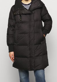 Svart pufferjacka med hög krage, horisontell quiltstruktur och framfickor. Har dragkedja och tryckknappsstängning. Texturerad tyg.