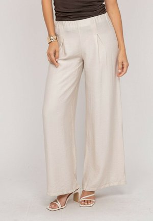Femme portant un pantalon ample beige clair à jambes larges avec des plis, associé à des sandales blanches à talons et un haut foncé, debout devant un fond uni.