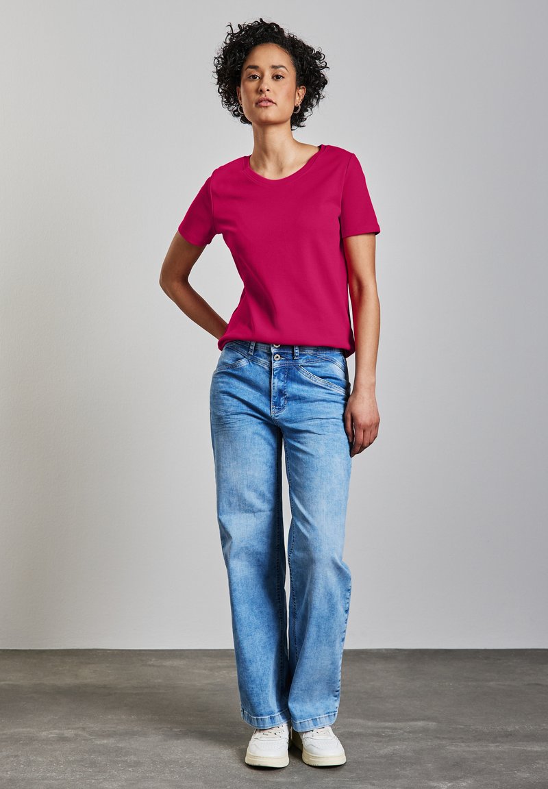 Fuchsia kortärmad t-shirt med rund halsringning, kombinerad med ljusblå vida jeans och vita sneakers, mot en neutral bakgrund.
