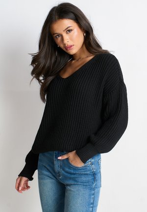 Young woman with long dark hair wearing a black ribbed sweater and blue jeans, posing with one hand in pocket against white background.