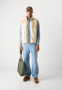 Beige fleece vest with a tan pocket, light blue denim shirt, light blue jeans, brown suede shoes, and an olive green tote bag with black straps.