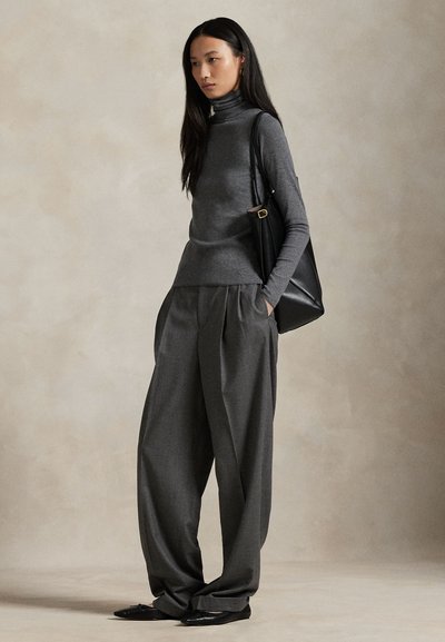 Naine, kellel on seljas hall turniirkrae, laiade säärekatetega hallid püksid, mustad madalad kingad ja kandes musta õlakotti ühevärvilise beeži tausta ees.