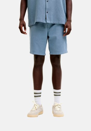 Persona con pantalones cortos azul claro y camiseta a juego, calcetines blancos con rayas verdes y zapatillas beige, de pie contra un fondo liso.