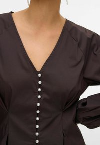 Dunkelbraune Bluse mit tiefem V-Ausschnitt, Perlenknopfdetails auf der Vorderseite und puffärmeln. Weicher Stoff mit strukturierter Passform.