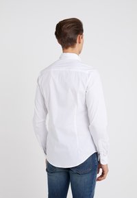 Bílá košile s dlouhým rukávem, rovného límce a knoflíkovými manžetami, s hladkou texturou a zakřiveným lemem, zkombinovaná s modrými džíny.