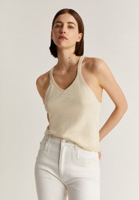 Débardeur en tricot de couleur crème avec un décolleté en V et des détails en chevron subtils, associé à un jean blanc taille haute. Texture lisse.