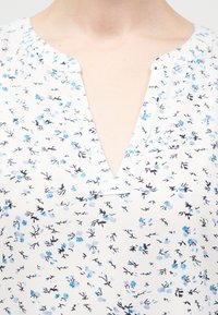 Λευκό πουκάμισο με μικρό μπλε και μαύρο ανθικό σχέδιο και V-σχήμα στο λαιμό σε ένα άτομο.