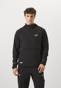 Μαύρο αδιάβροχο μπουφάν με φερμουάρ μπροστά, κουκούλα και ανακλαστικές λεπτομέρειες. Διαθέτει λογότυπο στο στήθος και στοιχεία εξαερισμού.